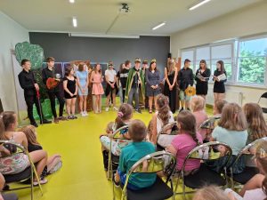 Uczniowie klasy siódmej biorący udział w przedstawieniu pozują do wspólnego zdjęcia stojąc w jednym rzędzie na sceną. Przed nimi na krzesełkach siedzą uczniowie klas 0- III.