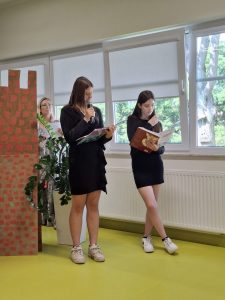 Dwie uczennice klasy VII w czarnych sukienkach stoją obok siebie. Dziewczynki trzymają w dłoniach mikrofony oraz otwarte książki, z których czytają tekst.