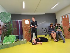 Scena z przedstawienia przygotowanego przez klasę siódmą. Nad leżącą na podłodze księżniczką stoi uczeń ubrany na czarno, który trzyma w dłoni długi miecz. W tle widać króla i królową siedzących na krzesłach z opuszczonymi w dół głowami oraz dekorację przedstawienia.
