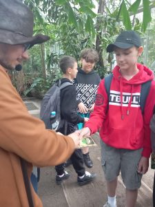 Uczeń wyciąga kartę, w stronę przewodnika, który przystawia mu pieczątkę. W tle widać stojących dwóch uczniów oraz palmy.