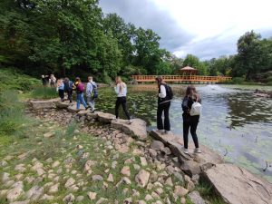 Uczniowie idą rzędem po dużych kamieniach ułożonych wzdłuż brzegu stawu.