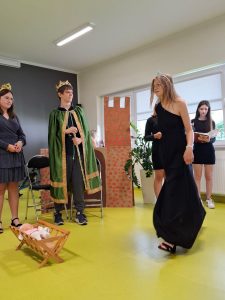 Zdjęcie wykonane podczas przedstawienia klasy siódmej. Na pierwszym planie widoczna jest idąca w czarnej sukni uczennica. Przed nią na podłodze stoi kołyska z lalką, za którą stoi dwóch uczniów w przebraniu króla i królowej.