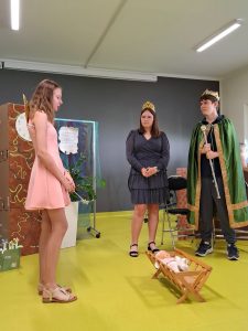 Uczennica klasy siódmej ubrana w jasnoróżową sukienkę i z przyczepionymi na plecach skrzydłami wróżki stoi nad kołyską z lalką. Obok stoi dwóch innych uczniów w strojach króla i królowej.