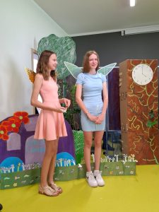 Dwie uczennice klasy siódmej stoją obok siebie. Dziewczynki mają na plecach przyczepione skrzydła wróżek i są ubrane w sukienki w pastelowych kolorach. W tle widać dekorację przedstawienia.