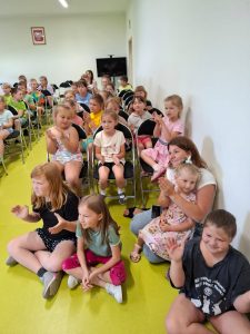 Uczniowie klas 0 - III oglądający występ klaszczą. Część uczniów siedzi w siadzie skrzyżnym na podłodze. Reszta widowni siedzi na krzesełkach ustawionych w rzędy.