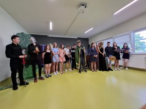 Uczniowie klasy siódmej biorący udział w przedstawieniu stoją w jednym rzędzie. Jeden z uczniów ma na głowie hełm, a stojący obok niego kolega podpiera się o długi miecz.