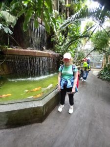 Uczennica stoi w palmiarni przy sztucznym wodospadzie u podnóża, którego pływają duże ryby.