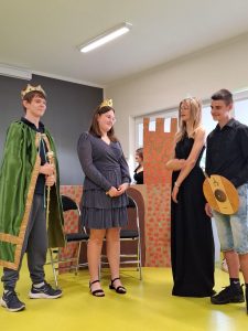 Czworo uczniów klasy siódmej odgrywa scenę. Dwoje uczniów przebranych jest za króla i królową, a na przeciwko nich stoi dwójka uczniów odgrywających role księżniczki i księcia.