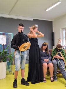 Uczniowie grający księcia i księżniczkę stoją obok siebie. Księżniczka ubrana w czarną długą suknię poprawie dłońmi włosy. Stojący obok książę trzyma w dłoni tarcze i zabawkowe czarnego konika.