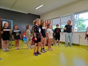 Uczniowie klasy drugiej stoją w dwóch rzędach. Część z nich trzyma w dłoniach lub ma przyczepione do bluzki tabliczki z angielskimi napisami. Nauczycielka stojąca z boku trzyma w dłoni mikrofon, który przysuwa do ucznia stojącego obok niej.