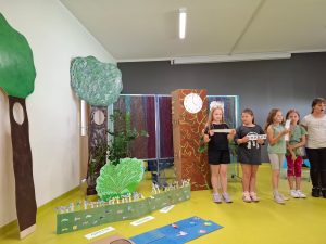 Na zdjęciu widoczne są uczennice, które trzymają wąskie karteczki z napisami. Obok nich stoi dekoracja - wycięte z papiery drzewa oraz trawnik.