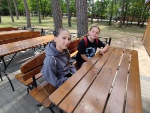 Dwie uczennice siedzą przy drewnianym stole i lekko się uśmiechając spoglądając w kierunku obiektywu.