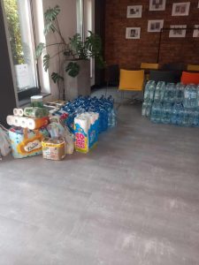 Zdjęcia sali z szara podłogą, na której ustawione są dary dla powodziań - ręczniki papierowe, butelki z wodą i pieluchy.