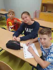 Troje uczniów siedzi przy stoliku i uśmiecha się do obiektywu. Jeden z chłopców podnosi w górę kciuk. Inny opiera dłonie o stół a pod nimi leży kartka papieru z przygotowanymi zadaniami.