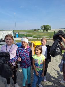 Cztery uczennice stoją na chodniku. Jedna z nich trzyma foliowy worek. Z boku widoczna jest dłoń podnosząca podobny worek w górę. Za dziewczynkami widoczne są plastikowe kosze na śmieci.