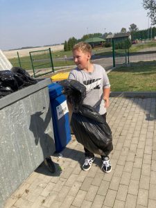 Uczeń klasy czwartej wrzuca czarny worek do kontenera na śmieci. Chłopiec spogląda w jego w stronę i się uśmiecha.