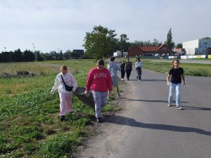 Uczniowie klasy szóstej idą asfaltową ścieżką. Dwie uczennice widoczne na pierwszym planie trzymają wspólnie czarny worek na śmieci. W tle widoczne są budynki szkoły.