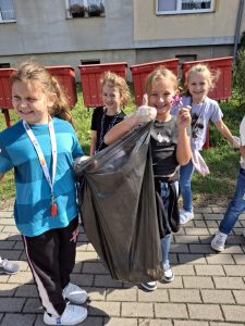 Cztery uczennice pozują do wspólnego zdjęcia. Wszystkie się szeroko uśmiechają. Dwie stojące z przodu trzymają w dłoniach, pomiędzy sobą czarny worek na śmieci. Jedna z dziewczynek podnosi w górę kciuki u obu dłoni.