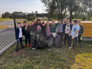 Klasa czwarta pozuje do wspólnego zdjęcia trzymając swoje ramiona oparte o kolegów. Przed nimi na trawniku leżą trzy czarne worki ze śmieciami.