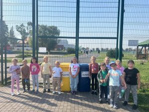 Uczniowie pozują do wspólnego zdjęcia stojąc przed bramą na boisko orlik. Za plecami dzieci widoczne są dwa plastikowe kosze na śmieci z napisami "PAPIER", "PLASTIK".