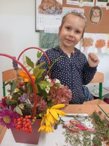 Uczennica siedzi przy stoliku na, którym ustawiony jest przygotowany przez nią jesienny koszyk. Dziewczynka patrzy przed siebie i się uśmiecha trzymając jedną rękę podniesioną na wysokość barków.