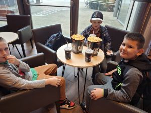 Trzech uczniów siedzi w fotelach przy okrągłym stole. Chłopcy spoglądają w kierunku obiektywu a przed nimi na stole leżą pudełka z popcornem i papierowe kubki z napojami.