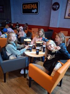 Cztery dziewczynki siedzą w fotelach przy okrągłym stole. Przed nimi na stoliku ustawione są pudełka z popcornem i papierowe kubki z napojami. Dziewczynka widoczna na pierwszym planie uśmiecha się przechylając głowę i podnosząc dłoń z wyprostowanymi dwoma palcami układającymi się w literę V.