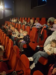 Uczniowie siedzą w dwóch rzęcha na sali kinowej. Dzieci trzymają pudełka z popcornem i patrzą przed siebie.
