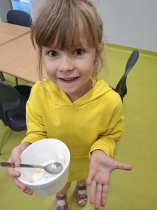 Uczennica oddziału przedszkolnego stoi z pustą miska w dłoni. Dziewczynka się uśmiecha i wyciąga dłoń w geście pokazującym, że miska jest pusta.