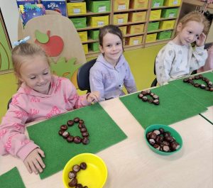 Trzy dziewczynki z oddziału przedszkolnego siedzą obok siebie przy stole. Przed nimi leżą zielone filcowe podkładki, na których ułożone są korale z kasztanów. Przed podkładkami na stole ustawione są miseczki wypełnione kasztanami.