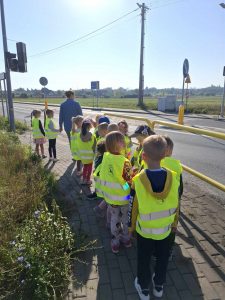 Uczniowie oddziału przedszkolnego idą parami po chodniku przy ulicy. Dzieci mają na sobie odblaskowe kamizelki.