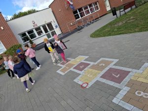 Uczniowie oddziału przedszkolnego stoją przy narysowanych na szkolnym boisku klasach. Jedna z dziewczynek wchodzi na pierwsze pole.