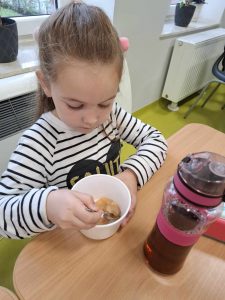 Uczennica oddziału przedszkolnego siedzi przy stoliku., Dziewczynka patrzy w miseczkę, która stoi przed nią. W ręku trzyma łyżeczkę umieszczoną w misce.