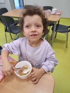 Uczennica oddziału przedszkolnego siedzi przy stoliku. Dziewczynka patrzy przed siebie z wysoko uniesioną głową. Przed nią stoi miseczka z jedzeniem, z której wyciąga łyżkę.