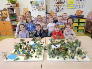 Uczniowie oddziału przedszkolnego pozują do wspólnego zdjęcia stojąc za złączonymi stołami, na których umieszczone są przygotowane przez nich platformy przedstawiające las. Dzieci do dekoracji użyły darów jesieni, przede wszystkim w postaci gałązek drzew iglastych.