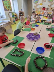 Zdjęcie uczniów oddziału przedszkolnego siedzących przy dużym stole. Przed dziećmi leżą zielone filcowe podkładki, na których dzieci tworzą korale z kasztanów.