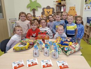 Uczniowie oddziału przedszkolnego pozują do wspólnego zdjęcia stojąc za dużym stołem na którym rozłożone są słodkie przekąski oraz upominki dla chłopców z okazji dnia chłopaka.