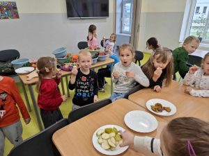 Uczniowie oddziału przedszkolnego siedzą lub stoją przy stołach. Dzieci jedzą kawałki owoców, które leżą na talerzach ustawionych na stole przed nimi. Na pierwszym planie widoczna jest uczennica sięgająca do talerza z gruszkami ustawionego na stole przed nią.