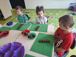 Troje uczniów oddziału przedszkolnego siedzi przy wspólnym stole. Przed dziećmi leżą zielone filcowe podkładki, na których ułożone są w rzędach kasztany. Przed podkładkami na stole leżą kolorowe miseczki wypełnione kasztanami. Jedna z dziewczynek szeroko otwiera buzię patrząc na kolegę siedzącego obok niej.