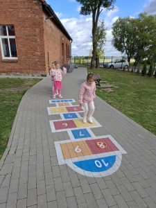 Dwie uczennice skaczą po kolorowych klasach namalowanych na chodniku szkolnego boiska.