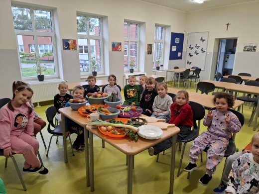 Uczniowie oddziału przedszkolnego siedzą na krzesełkach wokół stołu, na którym umieszczone są plastikowe miski i półmiski wypełnione warzywami i owocami. Dzieci uśmiechają się.