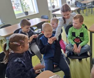 Uczniowie oddziału przedszkszolnego siedzą na krzesłach na szkolnej stołówce. Jedna z dziewczynek trzyma przy twarzy swojego kolegi kawałek buraka cukrowego. Chłopiec pochyla się nad kawałkiem i również trzyma go w dłoni.