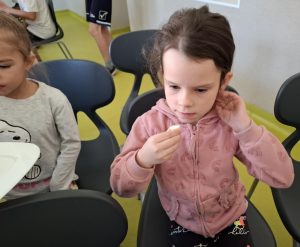 Uczennica oddziału przedszkolnego siedzi na krześle trzymając przed twarzą kawałek buraka cukrowego. Dziewczynka drugą dłonią poprawia swoje włosy na wysokości ucha.