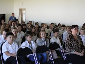 Zdjęcie uczniów siedzących na krzesłach podczas apelu z okazji Dnia Edukacji Narodowej. W pierwszym rzędzie widocznym na zdjęciu siedzi jedna z nauczycielek.