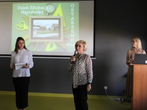 Na tle czarnej ściany, na której wyświetlona jest grafika z napisem "Dzień Edukacji Narodowej" oraz "OSTASZEWO 2024", stoją w odstępach od siebie od lewej strony: uczennica prowadząca apel, Pani dyrektor przemawiająca przez mikrofon, oraz nauczycielka języka polskiego nadzorująca przebieg uroczystości.