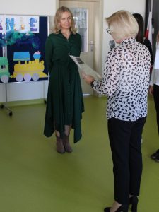 Nauczycielka w zielonej długiej sukience stoi na przeciwko Pani dyrektor trzymającej przed sobą dyplom, który za chwilę wręczy nauczycielce.