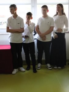 Czworo uczniów klasy piątej stoi obok siebie. Uczniowie ubrani są w białe koszule i czarne spodnie, a jedyna uczennica w grupie ma białą bluzkę i czarną długą spódnicę.