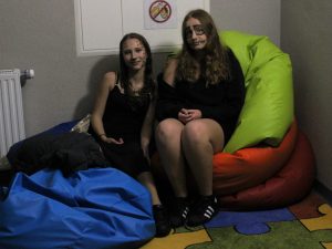 Dwie uczennice pozuje do wspólnego zdjęcia siedząc na kolorowych poduszkach. Dziewczynki ubrane są na czarno i mają pomalowane twarze czarną kredką.