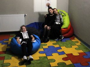 Trzy uczennice siedzą na kolorowych pufach na dywanie. Dwie z nich są obok siebie obejmując się. Jedna z nich trzyma w dłoniach replikę kosy. Dziewczynki się uśmiechają. Przed nimi na niebieskiej pufie siedzi ich koleżanka.