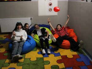 Cztery dziewczynki z klasy czwartej siedzą na kolorowych pufach ustawionych na dywanie. Dziewczynki uśmiechają się i pozują do wspólnego zdjęcia, Jedna z nich podnosi w górę szeroko rozstawione ręce nad którymi unosi się czerwony balon.